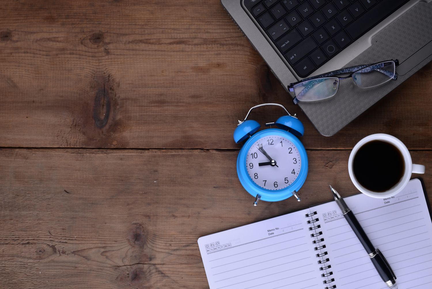 A notebook, clock, cup of coffee and laptop on a table