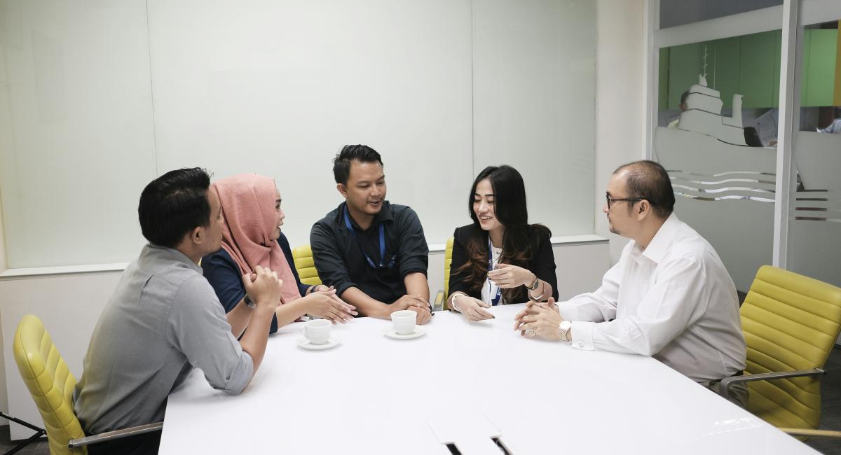 A group of five professionals sits around a white conference table, engaged in a team discussion in a modern office.